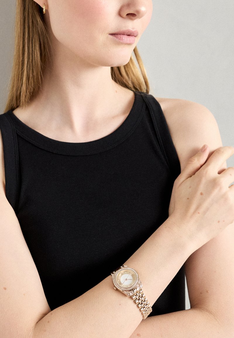 Montre-bracelet argent rond avec un cadran blanc, lunette ornée et bracelet en maillons métalliques ; portée sur un bras nu. Un débardeur noir est visible.