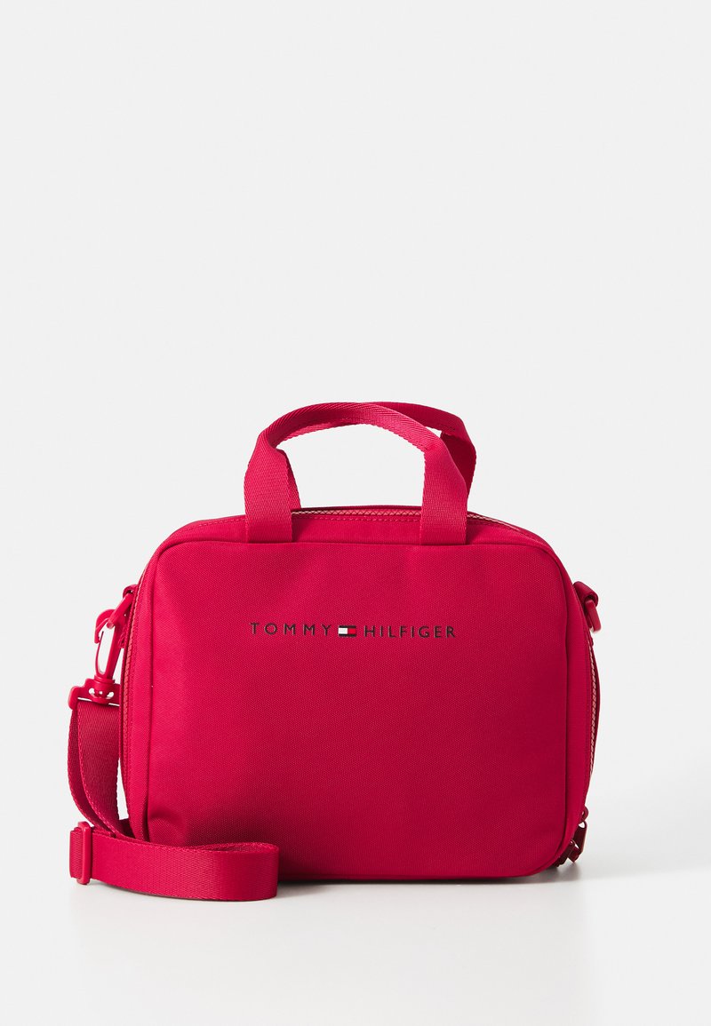 Sac en tissu rouge avec deux poignées et une bandoulière réglable, doté d'une fermeture éclair et du logo "TOMMY HILFIGER" en argent. Surface texturée.