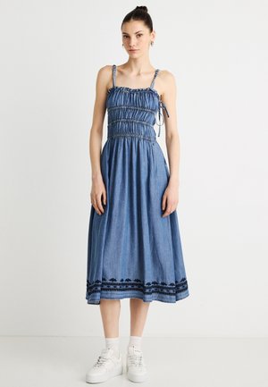 Jeanskleid mit gerafftem Oberteil, dünnen Trägern und einem fließenden Rock. Verziert mit schwarzer Stickerei am Saum. Kombiniert mit weißen Turnschuhen.