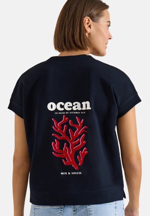 Kvinde iført mørkeblå kortærmet skjorte med rød koralgrafik og hvid tekst "ocean" på ryggen, kombineret med lyseblå jeans.