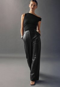 Fekete, egyvállas felsőt, magas derekú fekete bőrnadrágot és magassarkú szandált viselő nő, aki a zsebében tartja a kezét.