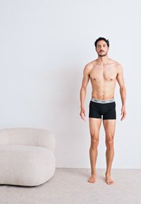 Fekete férfi boxer alsónadrág világosszürke elasztikus derékkal, amit egy álló férfi modell visel minimalista környezetben, egy ívelt krém színű széken.