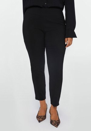 Kobieta w dopasowanych czarnych spodniach i szpilkach w panterkę z ostrym noskiem, stojąca na jednolitym tle.