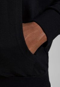 Black sweatshirt with ribbed cuffs and hem. Features a front kangaroo pocket. Close-up shows a hand resting in the pocket, displaying skin texture.