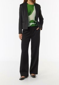 Musta räätälöity bleiseri vihreän ja valkoisen abstraktin kuvioidun topin päällä, yhdistettynä leveälahkeisiin mustiin housuihin ja mustiin balettitossuihin.