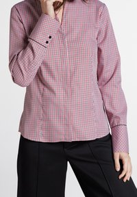 Camicia a quadri rossi e bianchi con scollo a V, maniche lunghe e bottoni neri a contrasto sui polsini. Texture liscia, vestibilità aderente.