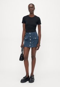 Svart t-skjorte med korte ermer, blå teksturert denim miniskjørt med frontknapper og lommer, kombinert med svarte kraftige sko og en liten veske.