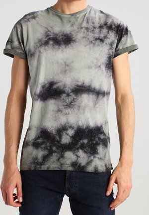 Persoon draagt een kortemouwen tie-dye t-shirt in zwart en grijs met donkere jeans, getoond van nek tot heupen met ontspannen armen langs de zij.