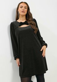 Schwarzes Samtkleid mit langen Ärmeln, das an der Halslinie mit einem Ausschnitt und einem Schleifendetail versehen ist, verziert mit kleinen weißen Punkten.