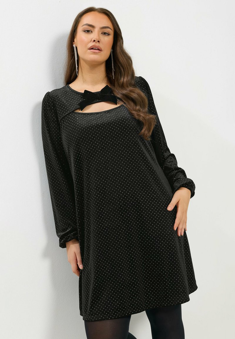 Schwarzes Samtkleid mit langen Ärmeln, das an der Halslinie mit einem Ausschnitt und einem Schleifendetail versehen ist, verziert mit kleinen weißen Punkten.