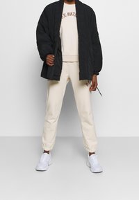 Vadderad svart jacka, krämfärgad sweatshirt med "P.E. NATION"-tryck, beigefärgade sweatpants och vita sneakers. Avslappnad lager-på-lager-outfit.