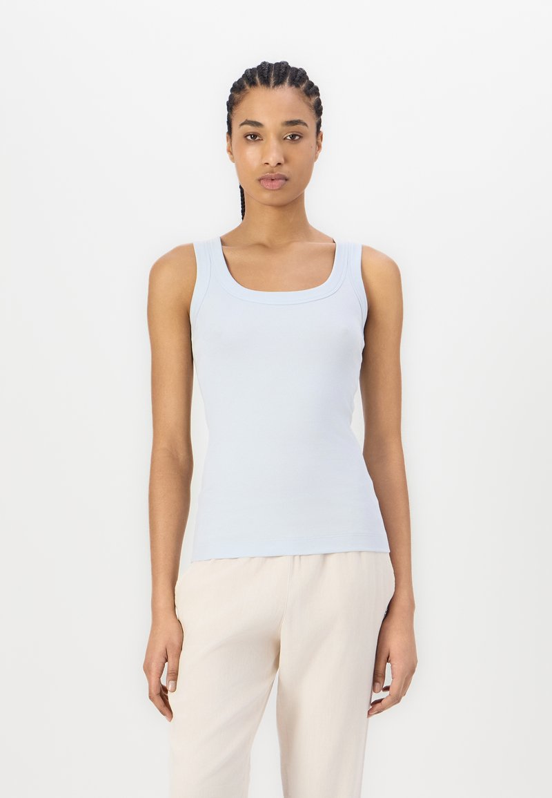 Lichtblauw tanktop met een vierkante halslijn, geribbelde textuur en brede schouderbanden, gecombineerd met beige losse broek tegen een effen achtergrond.