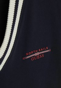 Tissu bleu marine avec bord côtelé rayé blanc et noir et logo rouge "NORTH SAILS GUESS" avec une corde et des étoiles brodées à l'avant.