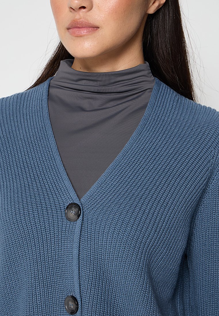 Blauer gestrickter Cardigan mit V-Ausschnitt, der aus strukturiertem Stoff besteht und zwei schwarzen Knöpfen hat. Über einem grauen Rollkragenoberteil getragen.