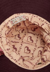 Chapéu balde de corduroy com um exterior em maroon. O interior apresenta um forro rosa com logótipos repetidos de "Barbour" e "FARM Rio" e acentos florais.