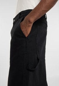 Pantalones de mezclilla negros con bolsillos laterales, una cintura resistente y detalles de costura visibles. El material parece duradero con una textura suave.