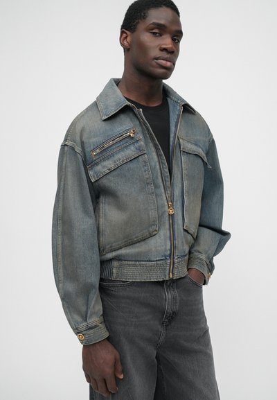 Denim jacket in faded blue with a zippered front, two chest pockets, gold hardware accents, and ribbed hem and cuffs, paired with dark jeans.