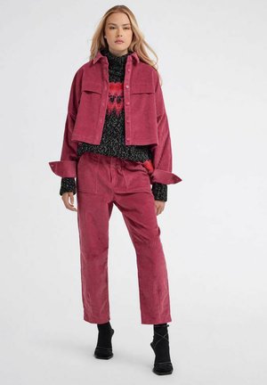 Donna che indossa una giacca corta in velluto a coste rosa e pantaloni abbinati sopra a un maglione nero e rosso con motivo, in piedi su uno sfondo bianco uniforme.