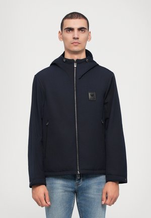 Veste marine avec capuche, fermeture éclair sur le devant et deux poches latérales. Présente un patch logo sur la poitrine et un tissu lisse et léger.