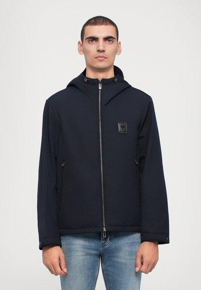 Marineblauwe jas met capuchon, rits aan de voorkant en twee zijzakken. Bevat een logopatch op de borst en heeft een gladde, lichte stoffen textuur.