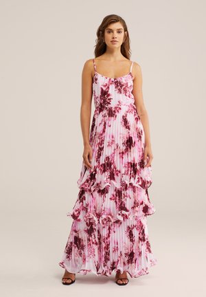 Młoda kobieta stoi ubrana w różowo-bordową, plisowaną suknię maxi na cienkich ramiączkach z falbanami i czarne sandały na obcasie z odkrytymi palcami.