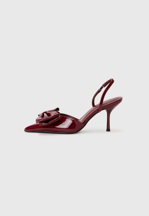 Chaussure à talon haut bordeaux brillant avec bride arrière, bout pointu et grand nœud décoratif à l'avant sur fond gris clair.