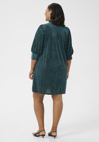 Tealfarbenes Samtkleid mit vertikalen Streifen, dreiviertellangen Puffärmeln und einem lockeren Schnitt, endet bis zur Mitte des Oberschenkels. Schwarze Riemchenabsätze runden das Outfit ab.