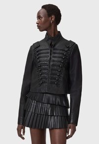 Zwarte leren militaire stijl jas met decoratieve lussen, hoge kraag en gestructureerde schouders, gecombineerd met een geplooide leren minirok.