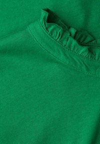 Zbliżenie zielonej tkaniny o gładkiej teksturze z falbanką przy dekolcie, podkreślające prostotę wzornictwa i żywy kolor.