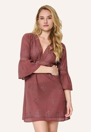 Blondínka s voľnými vlnitými vlasmi v mauve vyšívaných šatách s zvonovými rukávmi a výstrihom do V na jednoduchom pozadí.