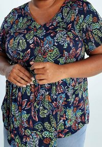 Femme portant un chemisier à motifs floraux bleu foncé à manches courtes, les mains superposées au niveau de la taille.