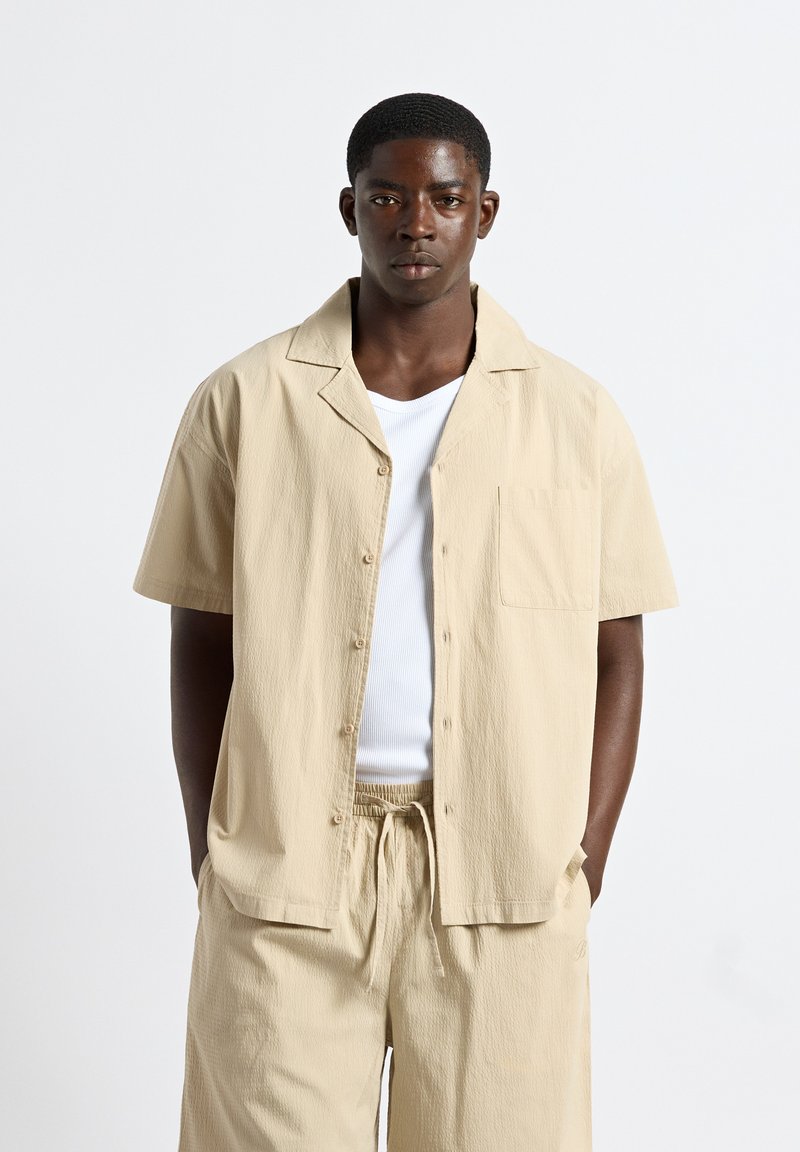 Jeune homme portant une chemise beige à manches courtes boutonnée et un short à cordon assorti par-dessus un débardeur blanc, debout les mains dans les poches.