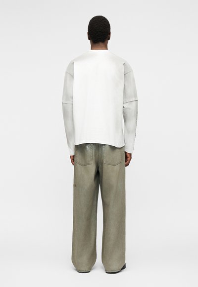 Άνδρας που φοράει υπερμεγέθη ξεβάμμενο γκρι τζιν και λευκό πουκάμισο με μακριά μανίκια, στέκεται στραμμένος με την πλάτη σε ένα απλό ανοιχτό φόντο.