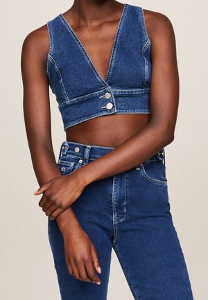 Vrouw draagt een mouwloos denim crop top met twee knopen aan de voorkant en hooggetailleerde denim jeans, armen gekruist ter hoogte van de taille.