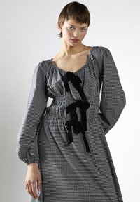 Joven con un vestido de cuadros en blanco y negro, con mangas largas y abullonadas, y lazos de terciopelo negro, de pie contra un fondo claro y uniforme.