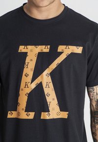 Černé bavlněné tričko s velkým oranžovým písmenem "K", které je zdobeno menšími vzory loga v opakovaném designu. Klasický kulatý výstřih.