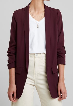 Woman wearing a burgundy blazer over a white v-neck shirt and cream high-waist pants, accessorized with layered silver necklaces.