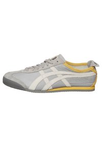 Šedá sportovní obuv vyrobená z hladké a strukturované kůže s bílými akcenty, žlutým lemováním a gumovou podešví. Obsahuje šest dírek na šněrování a prošívaný design.