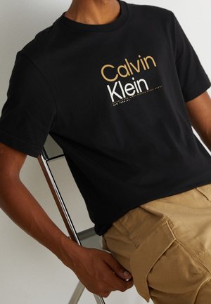 Person iført sort Calvin Klein t-shirt og beige cargobukser, siddende på en metalstol mod en ensfarvet baggrund.