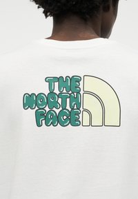 Άτομο με σκούρα σγουρά μαλλιά φορεί λευκό πουκάμισο με πράσινο κείμενο «THE NORTH FACE» και μπεζ αφηρημένη ημικυκλική λογότυπο στο πίσω μέρος.