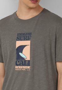 Graues T-Shirt mit kurzen Ärmeln, das einen grafischen Druck in Blau- und Pfirsichtönen zeigt, mit dem Text "ENDLESS SURF" und einem stilisierten Wellenmuster.