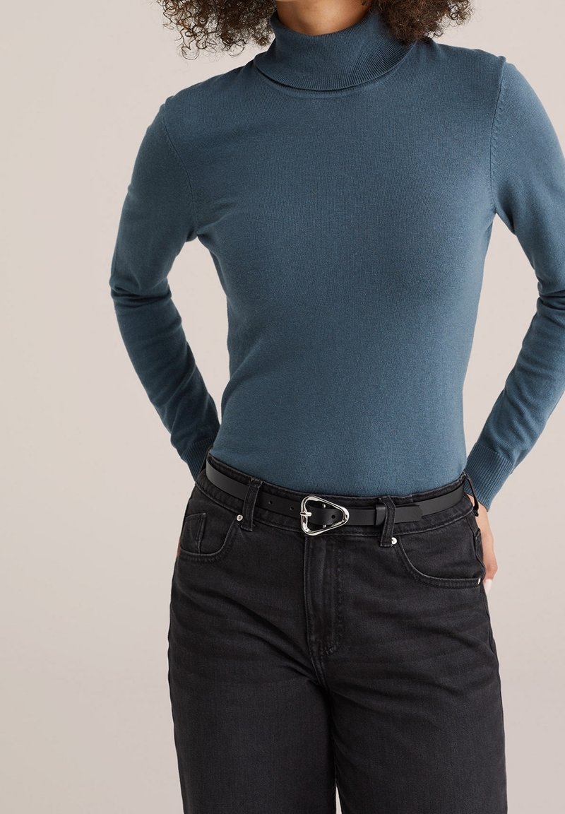 Femme portant un pull à col roulé bleu sarcelle rentré dans un jean noir taille haute avec une ceinture noire ornée d'une boucle triangulaire argentée.