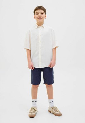 Chemise blanche à manches courtes à boutons, shorts bleu marine, baskets beiges et chaussettes blanches avec inscriptions noires, se tenant les mains sur les côtés devant un fond blanc.