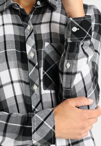 Black and white plaid button-up shirt, featuring a pocket, button cuffs, and a soft fabric texture with overlapping check patterns.