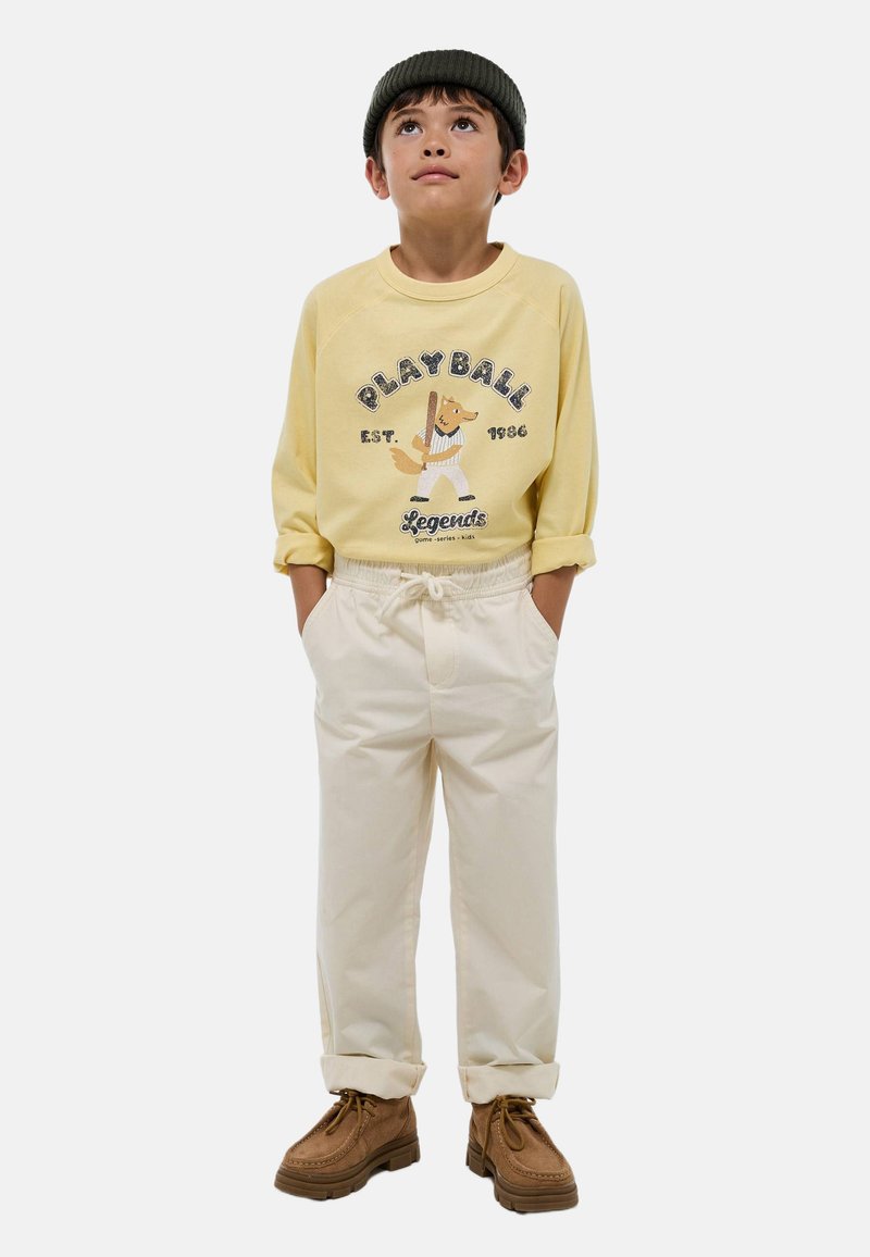 Sweatshirt jaune avec un graphisme "PLAYBALL" et un mascotte de dessin animé, pantalon élastique crème à taille, et chaussures brunes à lacets. Le modèle porte un bonnet noir.