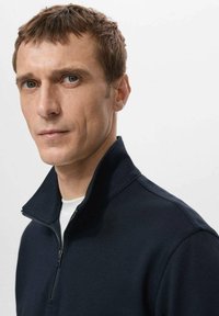 Tmavomodrá mikina na zips s golierom vyrobená z hladkej tkaniny, s kontrastným bielym okrúhlym výstrihom pod ňou a minimalistickým dizajnom.