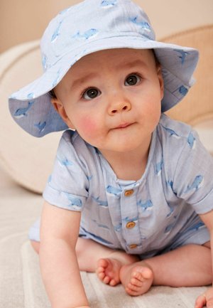 Vauva, joka käyttää sinisin valaskuvioidun haalarin ja siihen sopivan aurinkohatun, istuu vaalealla pinnalla ja katsoo ylös laajoin silmin.