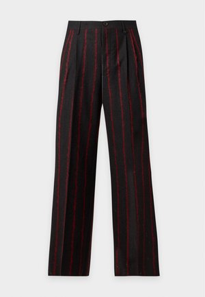 Homme debout portant un sweat à capuche noir avec un logo, un pantalon noir ample avec des rayures verticales rouges, des chaussures noires, sur un fond clair uni.