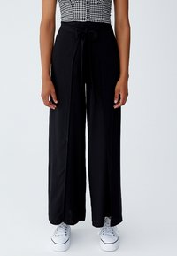 Zwarte wijde broek met een strikband, soepel materiaal en voorzakken. Gecombineerd met witte sneakers, wat een casual stijl laat zien.