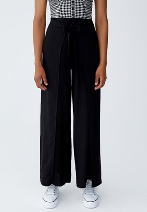 Pantalons larges noirs avec une ceinture nouée, tissu lisse et poches avant. Associés à des baskets blanches, mettant en avant un style décontracté.